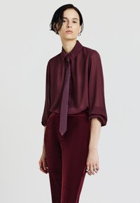 Blusa trasparente bordeaux con colletto e cravatta lunga coordinata, abbinata a pantaloni attillati in velluto bordeaux. Texture liscia, vestibilità rilassata e dettagli con bottoni.