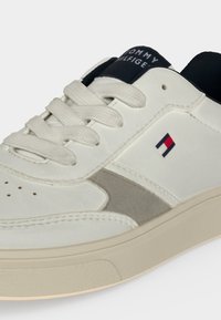 Λευκό και γκρι sneaker Tommy Hilfiger με λευκά κορδόνια, λογότυπο στο πλάι και μαύρη ετικέτα στη γλώσσα σε φωτεινό φόντο.