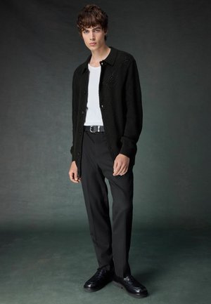 Sort tekstureret strikket cardigan over en hvid t-shirt, parret med sorte bukser og sorte sko. Outfittet har en knappet lukning og en tætsiddende stil.