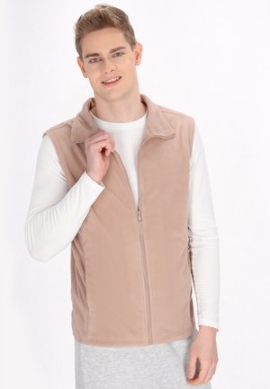 Beige színű bolyhos mellény, cipzárral elöl és gallérral, fehér hosszúujjú ing fölött viselva. Simasága és minták hiánya jellemzi.