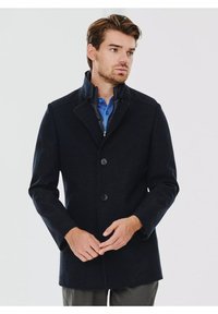 Czarny płaszcz z wełny, z wysokim kołnierzem, trzema guzikami, wewnętrznym zamkiem oraz teksturowanym wzorem. Nosi się go na niebieskiej koszuli, z dopasowanymi rękawami.
