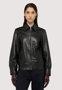 Černá kožená bunda s špičatým límcem, předním zipem a elastickými manžetami. Nosit s tmavými džíny a hnědými koženými rukavicemi.