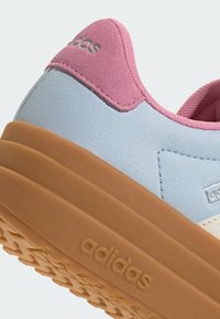 Gros plan sur le talon d'une basket avec un col en suède rose, une tige texturée bleu clair et une épaisse semelle en gomme marquée "adidas" sur le côté.