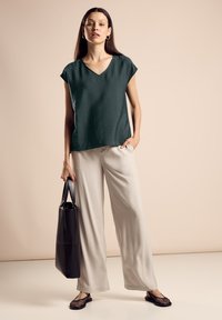 Mörkgrön v-ringad topp med korta ärmar, matchad med ljusbeiga vida byxor. Svart tote-väska och svarta nätstrumpor.