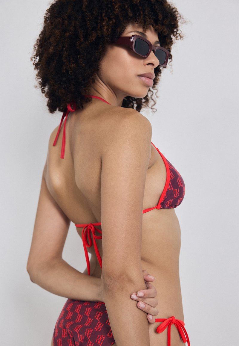 Raudonas bikini su pakartojančių logotipų raštu, su kaklo virvele ir užrišimu nugaros, derinamas su stačiakampiais saulės akiniais. Lygus, tamprus audinys.