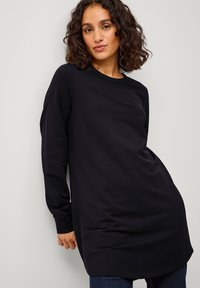 Zwarte lange mouw tuniek van zachte stof, met een ronde hals en een relaxte pasvorm, die voorbij de heupen valt, gecombineerd met donkere jeans.