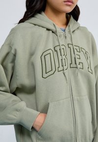 Green zip-up hoodie with "OBEY" in dark green lettering, featuring a cotton blend material, a kangaroo pocket, and ribbed cuffs.