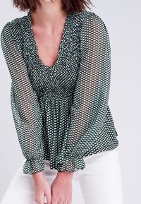 Blouse à motifs verts et noirs avec manches longues transparentes, corsage froncé et poignets froncés. Le design présente un décolleté en V profond.