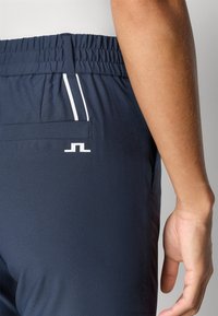 Jūrų pajėgų sportiniai šortai su elastinga juosmena, baltais akcento dryžiais, galiniu kišenėle ir logotipu šalia kišenės. Sklandus audinio paviršius.