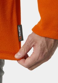 Pomarańczowy polar Polartec z metką u dołu. Materiał ma fakturę o miękkim, ciepłym wyglądzie; widoczna jest ręka trzymająca krawędź.