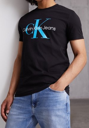Personne portant un t-shirt noir Calvin Klein Jeans avec un grand logo CK bleu et blanc, associé à un jean en denim bleu clair.