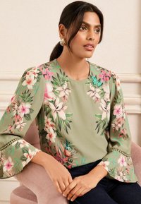 Floral blouse in olive green with bell sleeves. Features pink and white flowers, green leaves, and decorative trim along the sleeve edges.