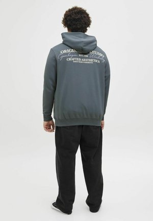 Dunkelgrauer Pullover-Hoodie mit einer Kordelzugkapuze, strukturiertem Stoff und weißem Drucktext auf dem Rücken. Kombiniert mit lockeren schwarzen Hosen.
