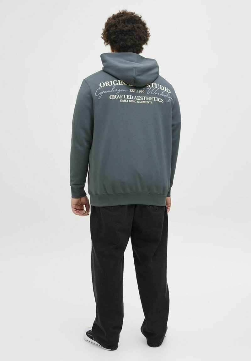 Donkergrijze pullover hoodie met een trekkoordcapuchon, gestructureerde stof en witte opdruk op de achterkant. In combinatie met losse zwarte broeken.