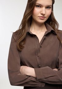 Camicia marrone con bottoni, colletto e maniche lunghe, caratterizzata da una consistenza morbida e una leggera lucentezza. Bottoni e cuciture visibili lungo la parte anteriore.