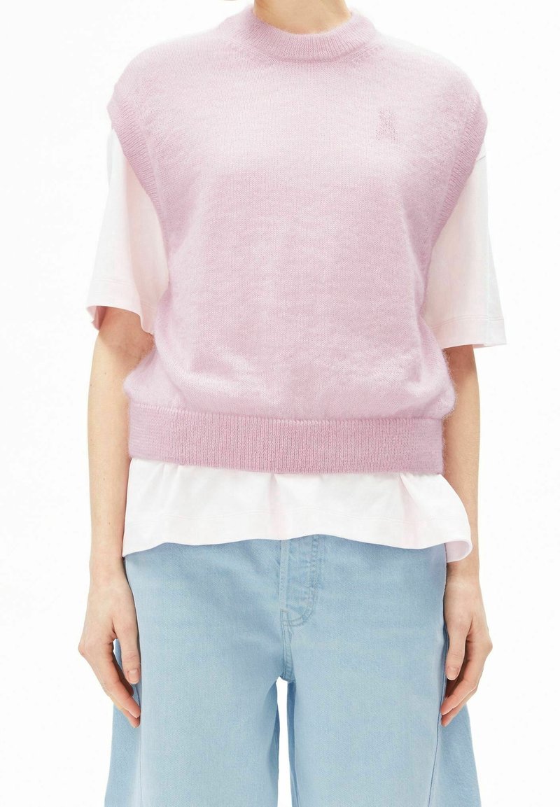 Personne portant un gilet en tricot rose sans manches sur une chemise blanche à manches courtes et un jean bleu clair à jambes larges.