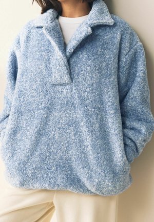 Persoon draagt een lichtblauwe, gestructureerde fleece trui met een brede kraag over een wit overhemd en beige broek, handen in de zakken.