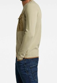 Beige Langarmhemd aus strukturiertem Stoff mit einer braunen Brusttasche. Kombiniert mit dunkelblauen Jeans mit Seitentaschen und einem Knopfdetail.