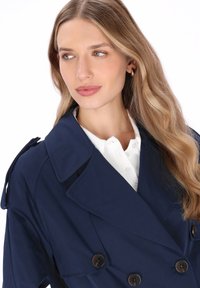 Marinblå dubbelknäppt trenchcoat med stora slag, markerade knappar och axelband, lagd över en vit skjorta med krage.