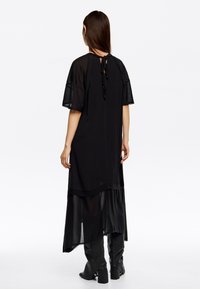 Mustat maksimekot, joissa on lyhyet, läpinäkyvät hihat ja kerrostettu helma. Takana on nauhakiinnitys ja sileä pinta.