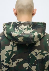 Kamuflāžas kapuci džemperis zaļos un brūnos toņos, ar regulējamu kapuci un izšūtu "Dickies" logo uz aizmugures. Mīksta auduma tekstūra.