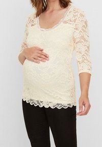 Lace maternity top in cream with three-quarter sleeves, featuring a scalloped hem and floral patterns, layered over a smooth fabric.