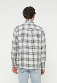 Καρό πουκάμισο με μακριά μανίκια σε λευκό και σκούρο μπλε, με άνετη εφαρμογή, μανσέτες με κουμπιά και λεπτομέρειες στο πίσω μέρος. Το ύφασμα φαίνεται υφασμένης υφής.