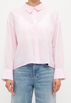 Lyserød skjorte med knapper og lange brede manchetter, båret sammen med blå jeans dekoreret med spredte hvide prikker.