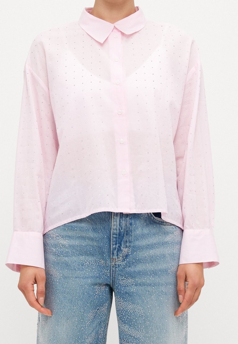 Chemise rose clair à boutons avec de longs poignets larges, portée avec un jean bleu orné de points blancs dispersés.