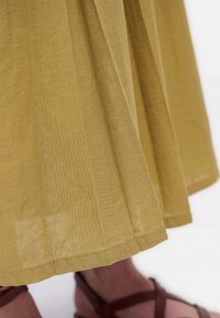 A mustard yellow skirt hem above brown strappy sandals on a white background.