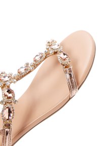 Sandalia plana con una tira brillante de color oro rosa, decorada con piedras de strass transparentes y rosas. Plantilla suave en beige con una ligera curva en la zona del dedo del pie.