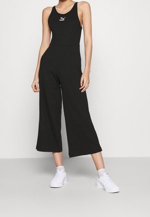 Vrouw draagt een mouwloze zwarte jumpsuit met een wit Puma-logo en witte Puma sneakers, staand tegen een effen lichte achtergrond.