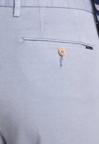 Pantalon bleu clair fabriqué dans un tissu à texture lisse, avec une poche arrière dotée d'un bouton doré et d'une petite étiquette de marque.