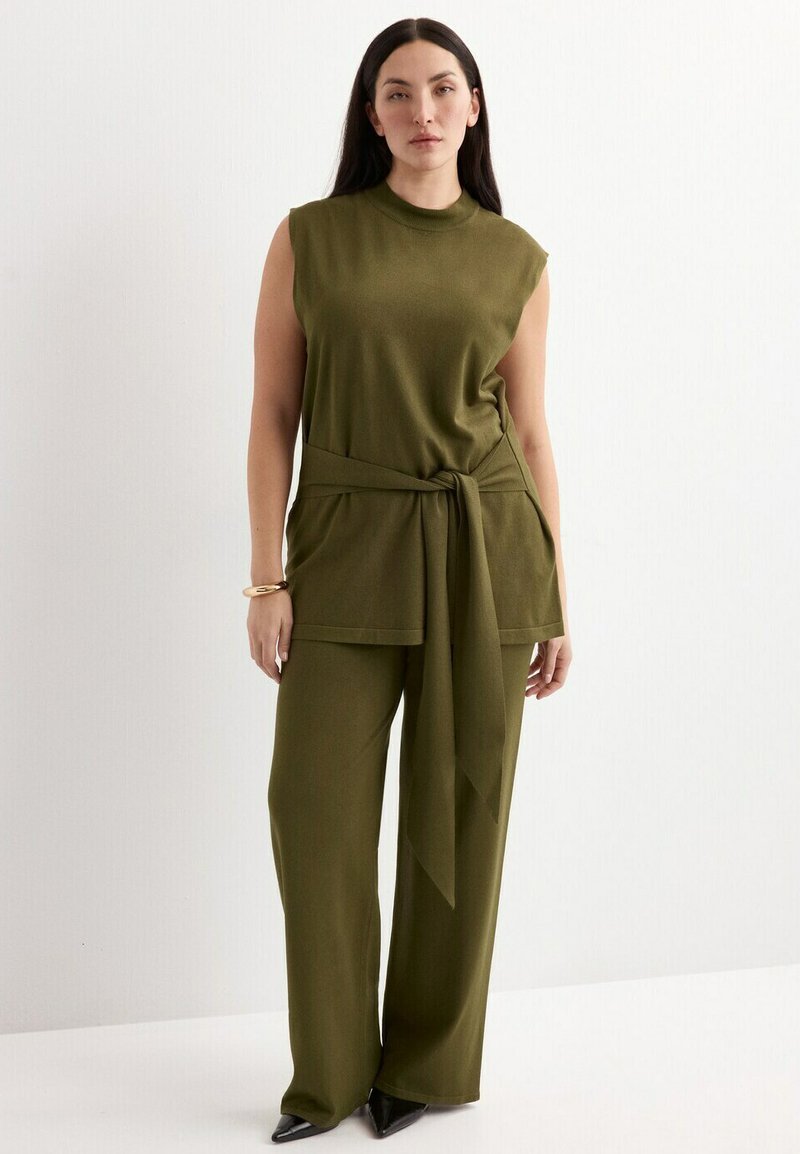 Femme debout portant un haut sans manches vert olive avec ceinture à la taille et un pantalon large assorti, chaussures noires à bouts pointus et bracelet en or.