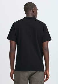 Mann in einem schlichten schwarzen Poloshirt mit kurzen Ärmeln und grauen Cargo-Hosen, der von hinten vor einem hellgrauen Hintergrund steht.