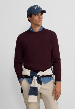 Suéter de punto burdeos sobre una camisa de botones azul claro, con un suéter a rayas crema y marino atado a la cintura y pantalones beige.