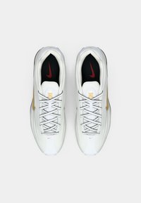 Par bijelih Nike tenisica s zlatnim swooshom i crnim ulošcima s crvenim logotipima, prikazano odozgo na bijeloj pozadini.