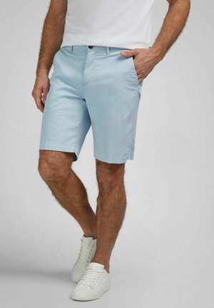 Man wearing light blue shorts, white sneakers, and a white shirt, standing with one hand in pocket on a gray background.