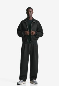 Mand står iført en sort zip-up jakke, sorte bredbenede bukser, sort skjorte og grå-hvide sneakers på en hvid baggrund.