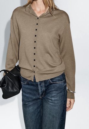 Woman wearing a brown buttoned long-sleeve top, blue jeans, holding a black leather bag in one hand.