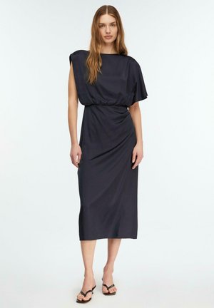 Kobieta ubrana w ciemnogranatową, bezrękawnikową sukienkę midi z marszczonym taliowaniem i asymetrycznym dołem, stojąca na tle białej ściany.