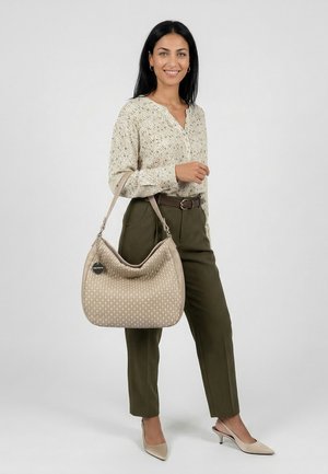 Vrouw glimlachend, gekleed in een beige blouse met patroon, olijfgroene broek, beige hakken, en een beige gestructureerde handtas met schouderriem.