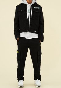 Svart denimjacka med silverdetaljer, framlädd fickor och en grön "GUESS"-lapp, tillsammans med svarta cargobyxor och vita sneakers.
