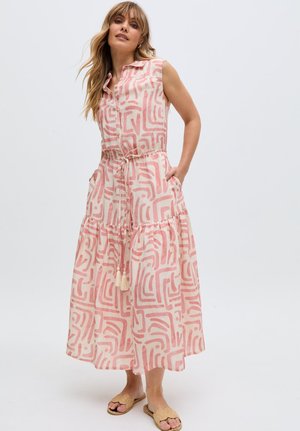 Frau trägt ärmelloses, knielanges Kleid mit Kragen, rosa abstraktem Muster und Tunnelzug an der Taille, steht mit Händen in den Taschen und beigen Sandalen.