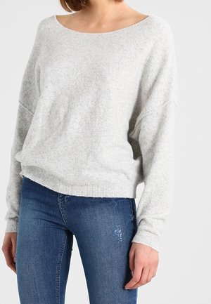 Femme portant un pull en maille gris clair à manches longues avec un col bateau et un jean bleu ajusté sur un fond uni.