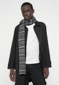 Ριγωτό γκρι κασκόλ από απαλό υλικό, με εναλλασσόμενα σκούρα και ανοιχτό γκρι σχέδια, φορεμένο πάνω από γκρι φούτερ και μαύρο μπουφάν.