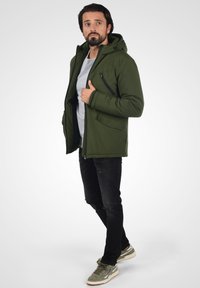 Grøn isoleret jakke med hætte, lynlås og åben front, to sidelommer, båret over en grå skjorte og sorte slidte jeans.