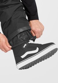 Sorte skate sko med hvide accenter, struktureret stof og gummisål; en hånd justerer det elastiske manchet på glatte sorte bukser.