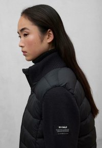 Chaqueta acolchada negra con cuello y mangas acanaladas. Presenta una textura de tejido de punto, cremallera lateral y una etiqueta Ecoalf visible.