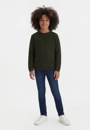 Niño con cabello rizado que lleva un suéter verde oscuro, vaqueros azules y zapatillas blancas, de pie frente a un fondo blanco liso.